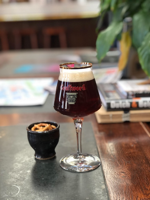 A dark sour beer in a tall-stemmed beer glass next to a brown egg cup full of pringles