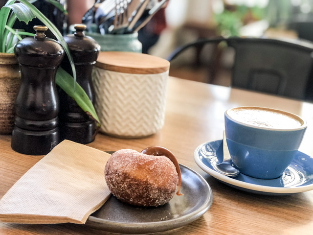 Flat White Coffee in blue cup and sauce and Doughnut on table with salt and pepper shaker, sugar bowl and small flax plant at Tees Street Cafe Oamaru
