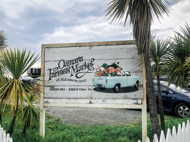 Wooden sign which reads "Oamaru Farmers Market, eat fresh with the locals, Harbour Area, Sundays 9:30am-1PM". There's also an illustration of a light blue flatbed truck packed with produce items.