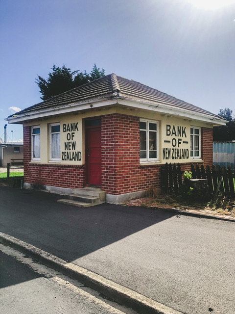 Small brick building with tiled roof and red door pictured from corner angle with Bank of New Zealand signs and white windows.