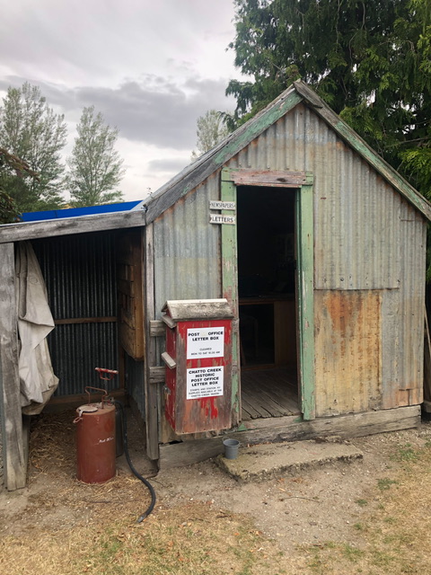 Old windowless post office formed of corrugated iron with a red post office attached to outside to left of door. Looks more like a shed than a post office.