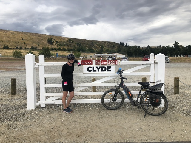 A young woman wearing cycling clothing and a helmet stands beside a sign on a white wooden gate which reads "Finish 04 Jan 2024, CLYDE". An ebike is parked next to the fence. In the background is a fairly empty carpark and brown grass hills.
