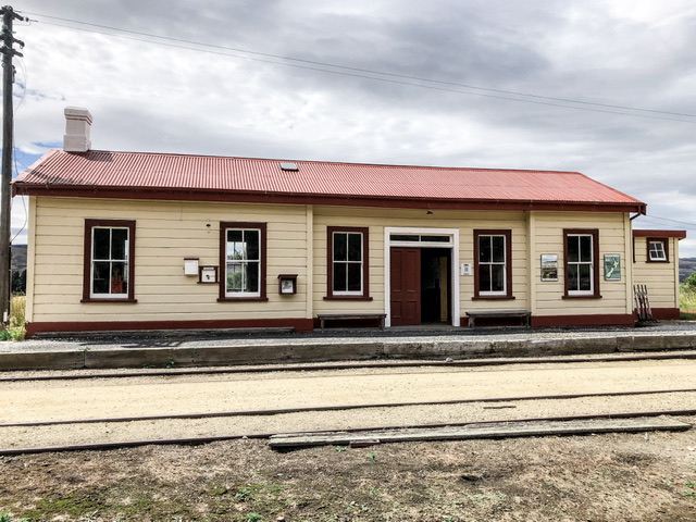 A creamy weatherboard former train station and platform with red detailing. There are five large windows and double doors.