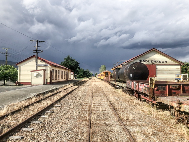 Middlemarch railway station with cream station buildings on each side of the tracks named with "Middlemarch" in dark red. An old yellow abandoned train is parked on the right track.
