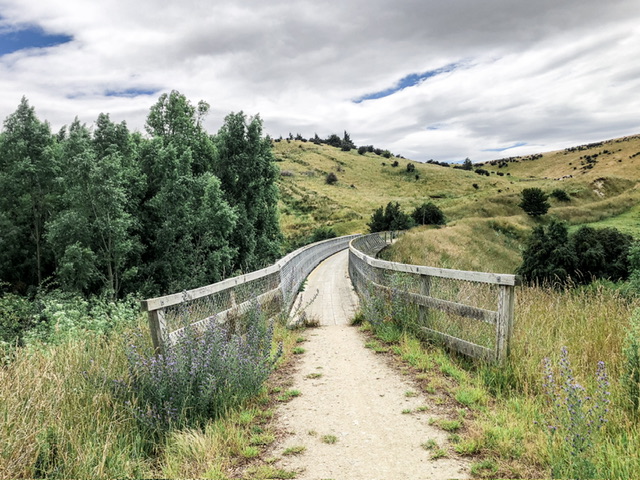 A wooden bridge curving to the right stretches into the distance between hills covered in dry grass. A group of tall green trees is to the left of the bridge.