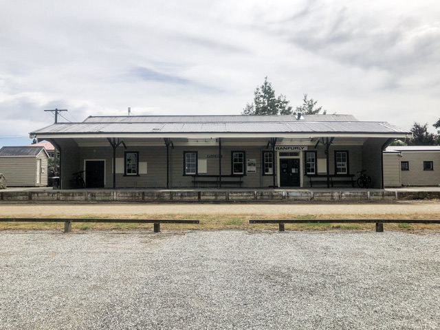 Exterior photo of historic Ranfurly railway station looking at the platform. The building is formed of grey weatherboards with a grey roof providing shelter on the platform, with black accents.