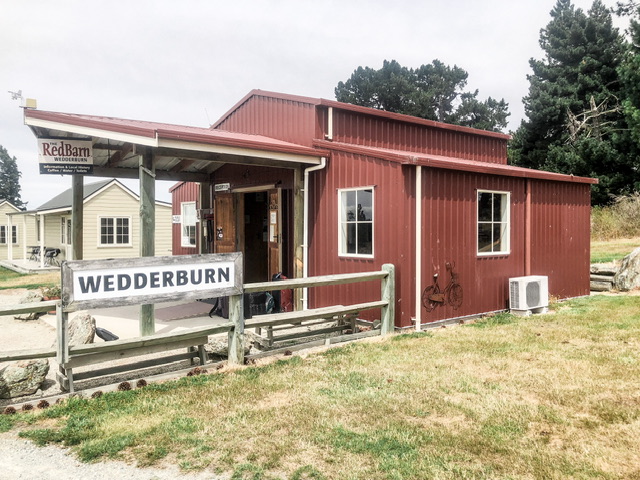 A modern looking red barn with a sign on fence outside which says "WEDDERBURN" and a hanging sign under shelter area which says "RedBarn".