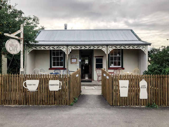 Exterior photo of The Kissing Gate Cafe Middlemarch. This is a converted white villa which has white signs on the fence advertising cups of tea, coffee, cold drinks and cappuccinos.