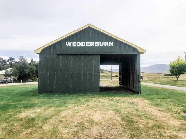 Green railway goods shed with "WEDDERBURN" painted on front. Surrounded by grass and you can see straight through the shed through open doors on the right.