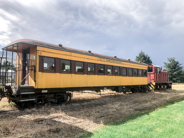 An old yellow wooden train carriage attached to a red train parked on grass on grey cloudy day. A sign on the train carriage says Taieri Gorge Railway.