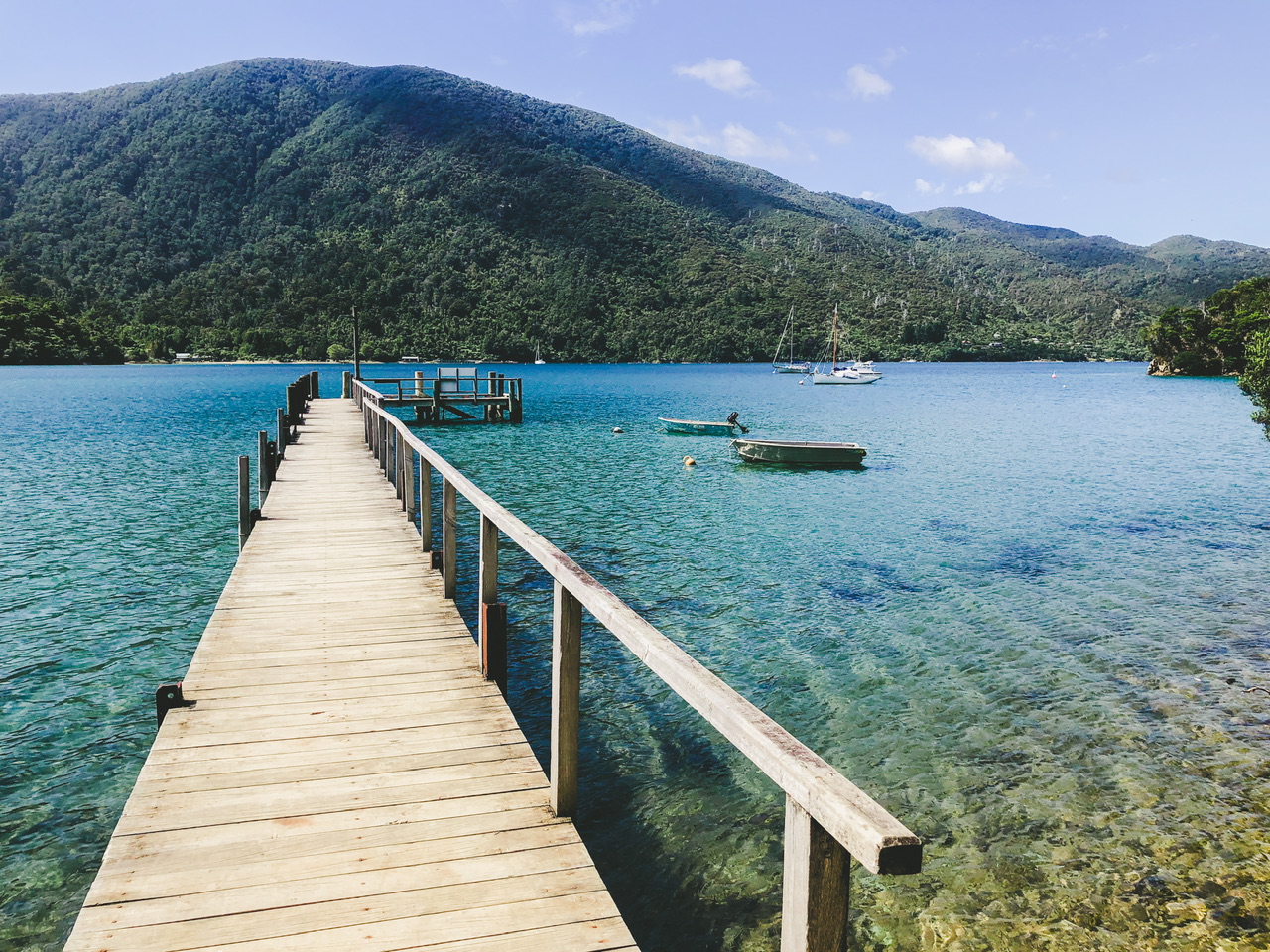 A wooden jetty juts out into Endeavour Inlet with a handrail on the right-hand side. The water is blue and clear and there ate two small boats close to the right side of the jetty. Small yachts and bush over the other side of the sound are visible in the background.