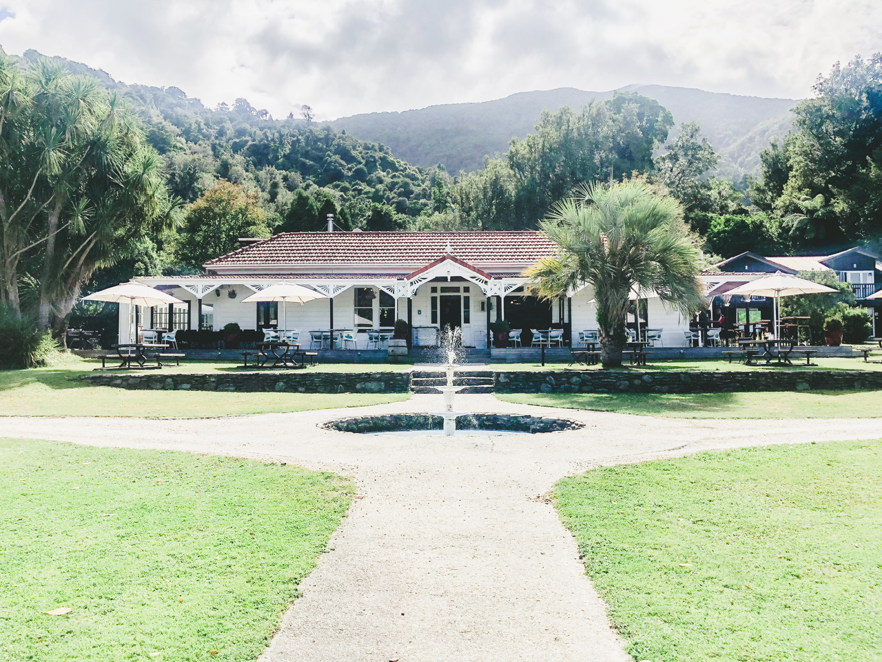 The white Furveaux Lodge with a fountain in centre fore-view and grass. Tables with sun umbrellas are set up in front of the lodge and on the balcony which wraps around.