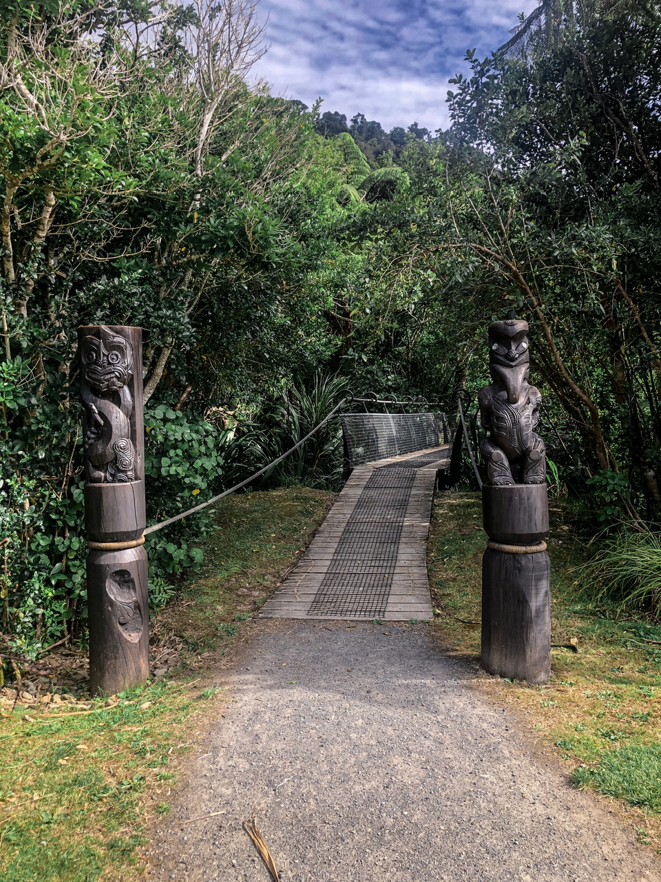 Two Māori totem carvings of around 6 feet in height stand with a path which leads to a footbridge running between them, through native New Zealand bush in Ship Cove.