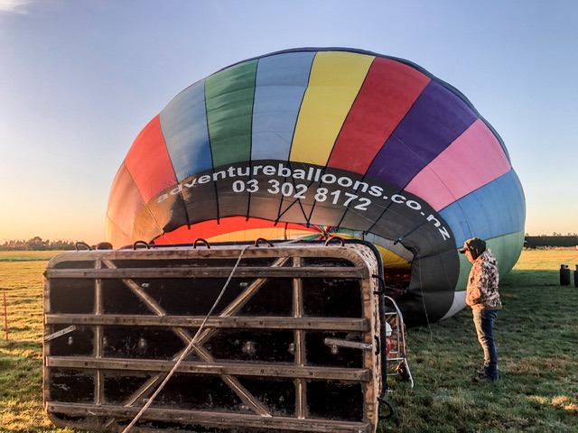 Colourful hot air balloon and basket lying on it’s side in a green paddock at dawn. The balloon has adventureballoons.co.nz URL on it and 03 302 8172. A man stands on the right of the balloon observing the inflation.