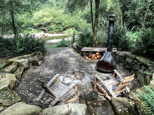 A fire pit with a chimney in a circular area surrounded by large stones. A river can be seen in the rear of the image. Two wooden chairs are in front of the firepit. Wood is stacked behind the enclosed firepit. Native bush is all around.