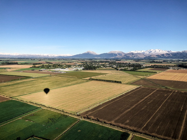 The silhouette of a hot air balloon flying over the Canterbury Planes with Methven and snow capped mountains in the distance. It's a cloudless blue morning.