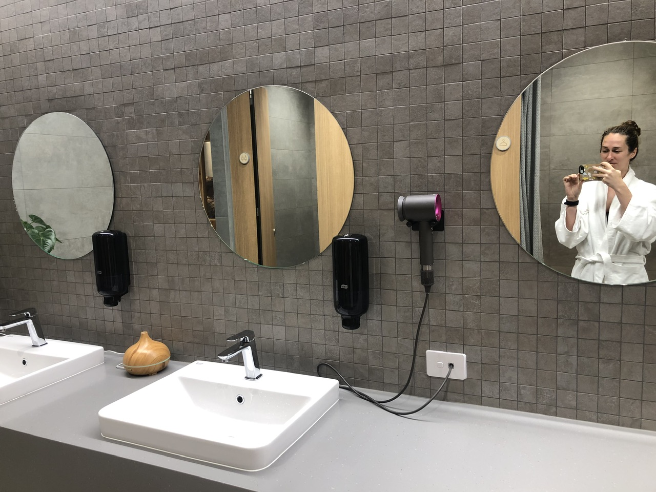 Young woman in white bathrobe takes a selfie in changing rooms at at Ōpuke Thermal Pools and Day Spa. A dyson hairdryer is on the grey tiled wall, there are also three mirrors, two soap dispensers and two sinks.