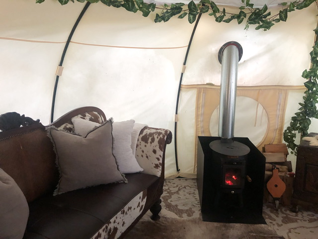 Interior of a glamping tent with an enclosed fire which is burning. On the left is a double couch covered in fake fur with two cushions on it, one is white and one grey. Fake ivy crawls over ceiling of glamping tent.