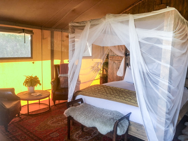 Four poster bed in Te Hāpua Coastal Glamping tent with two armchairs with a small coffee table between them on the left of the bed, a stool is at the foot of the bed.