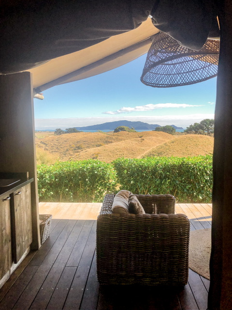 Wooden balcony with outdoor kitchen on left and single armchair on right, hanging light and tent structure above. Kāpiti Island is visible in the ocean in the distance behind green fields.