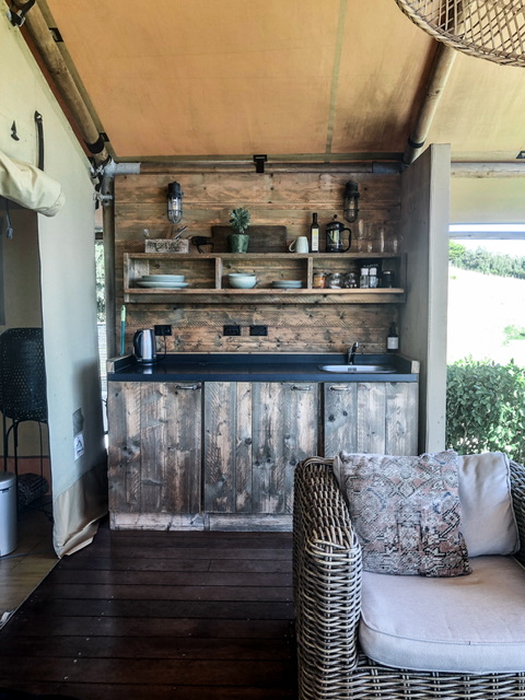 Compact country-style wooden Kitchen on a single wall with three cupboards at the bottom, bench with silver kettle on the left and sink on the right, and two shelves containing crockery and cooking tools above. A single cane armchair with white cushion is in the foreground.