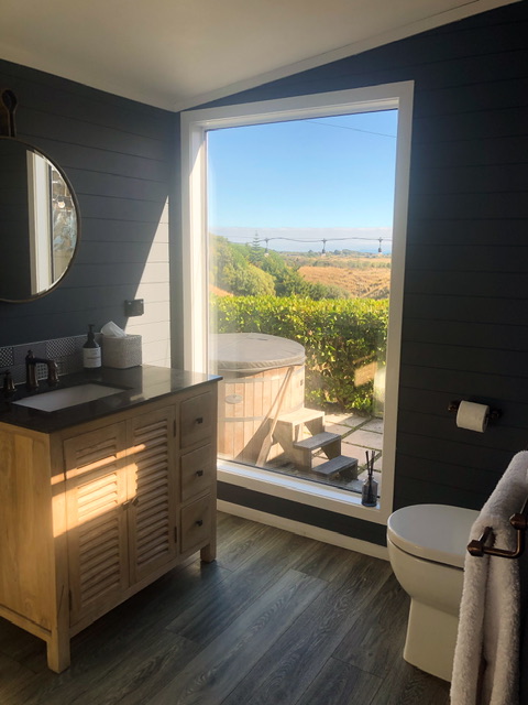 Bathroom at Te Hāpua Coastal Glamping with toilet, vanity and mirror. A large picture window looks our over a coverer round wooden hot pool and the ocean is visible in the distance behind green fields and bushes.