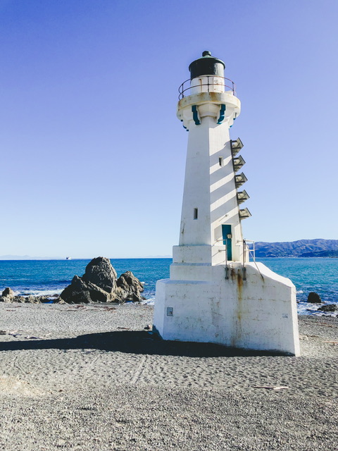 The white Pencarrow lower Lighthouse with rocky boulders to the left, blue ocean behind and Wellington's green hills over the other side of the harbour in the background.