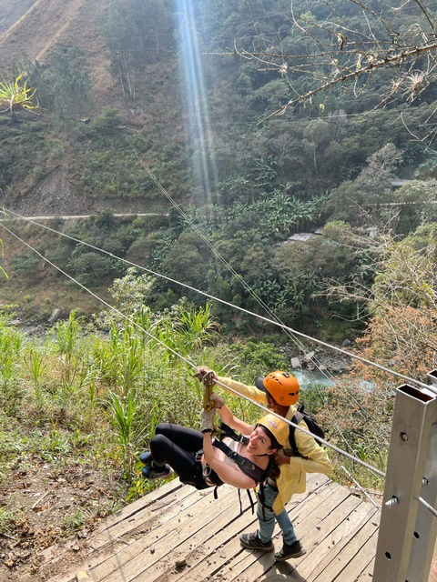 Young woman wearing black leggings and a strappy light pink singlet and yellow helmet smiles at camera whilst ziplining in Peru. A male in an orange helmet is assisting her to start the zipline over the jungle.0