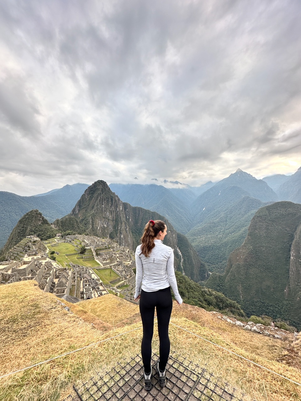 Young woman stands with her back to the camera looking slightly to the right over Machu Picchu. She has long brunette hair in a ponytail tied with a red scrunchie and is wearing black leggings, black and grey hiking boots and a cream coloured Lululemon Define Jacket.