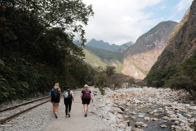 Three young women in activewear and backpacks walk with train tracks on their left and river on their right between Hidroeléctrica and Aguas Calientes in Peru. Mountain ranges are visible in the background of the image.