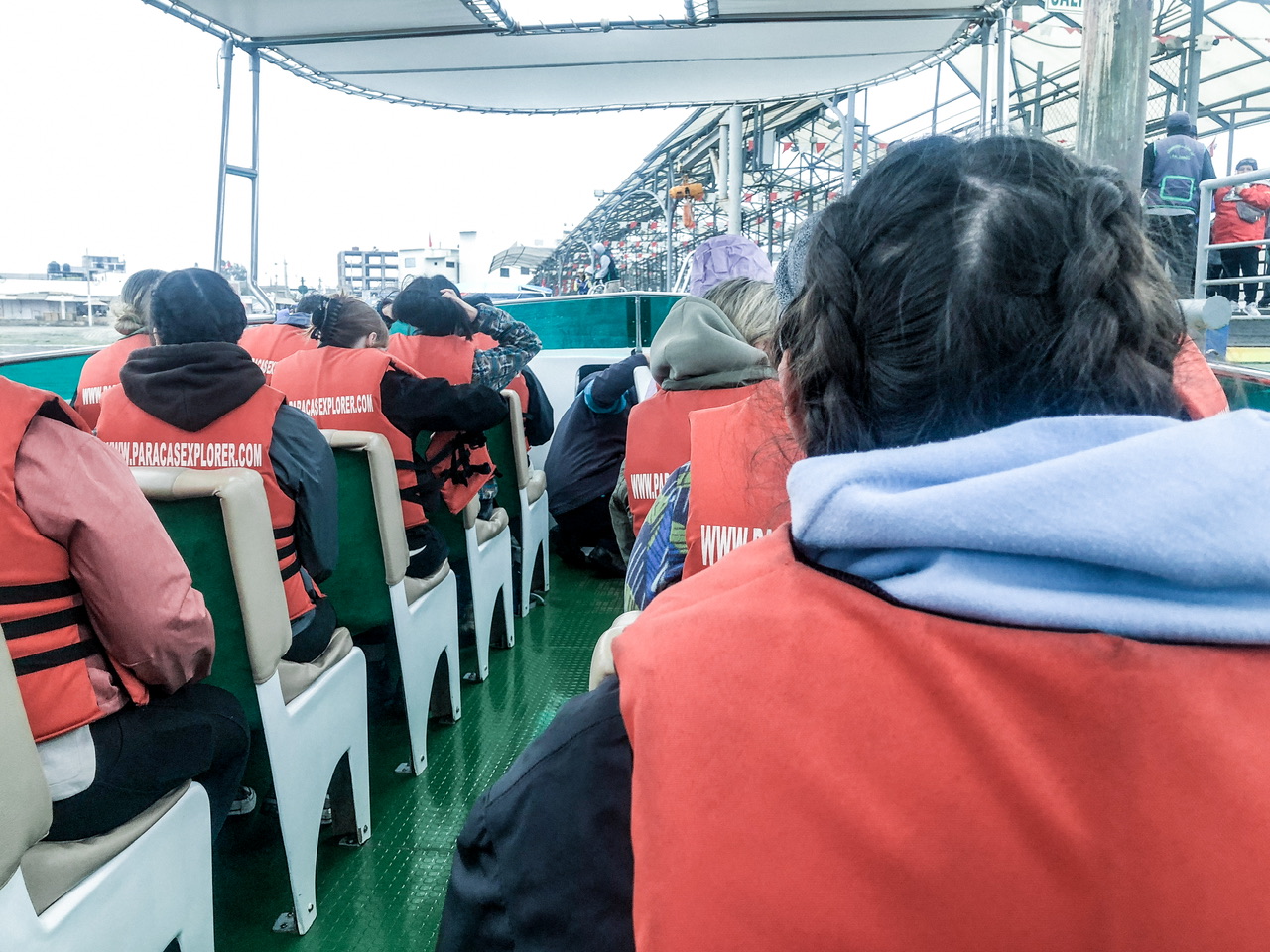 Large speed boat with wharf on right full of rows of people in red lifejackets, two people on each side of the boat.