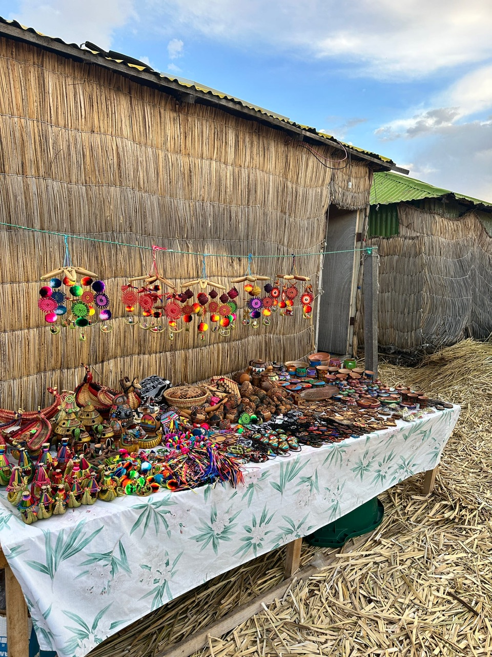 Colourful crafts for sale on a table covered in a simple white table cloth with a floral patter on a floating island on Lake Titikaka in Peru. The table is up against a thatched wall and the ground is covered in flax.