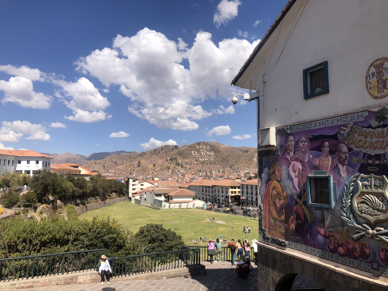 View in Cusco over mountains with "Viva El Peru" carved into the mountain. ON the right is a purple mural and straight ahead is a green fiend. Cusco city buildings are dotted around.