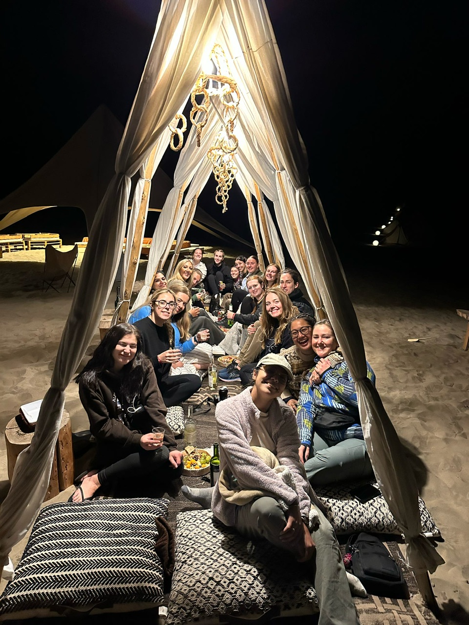 Eighteen young women dressed warmly and seated in two rows on cushions facing each other at desert glamping.