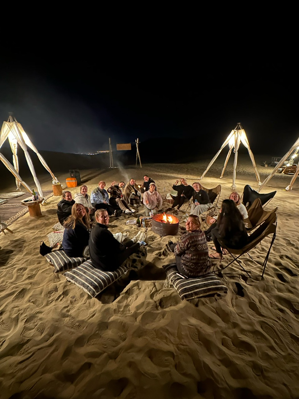 Fourteen young women dressed warmly and seated on cushions in a circle around a campfire.