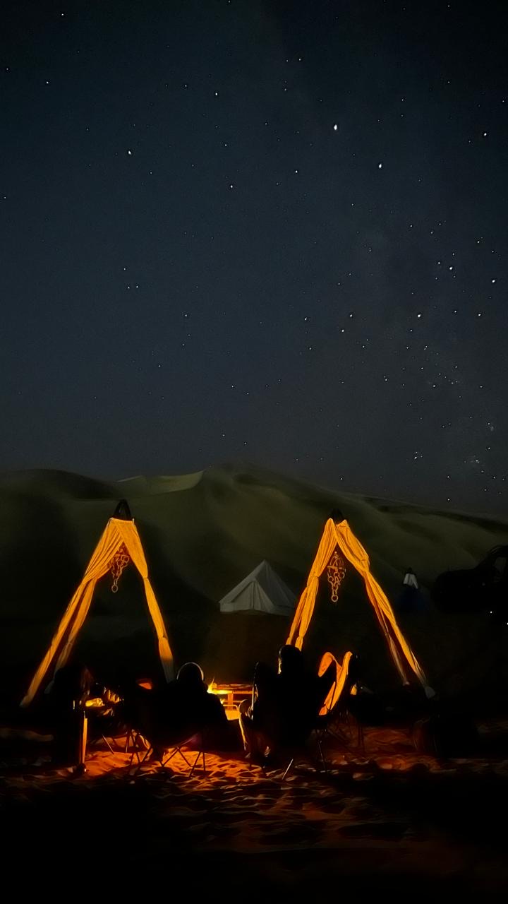 Starry sky over white glamping tents and two seated silhouette figures at Duna Camp Glamping site in the Hauacachina Desert in Peru.