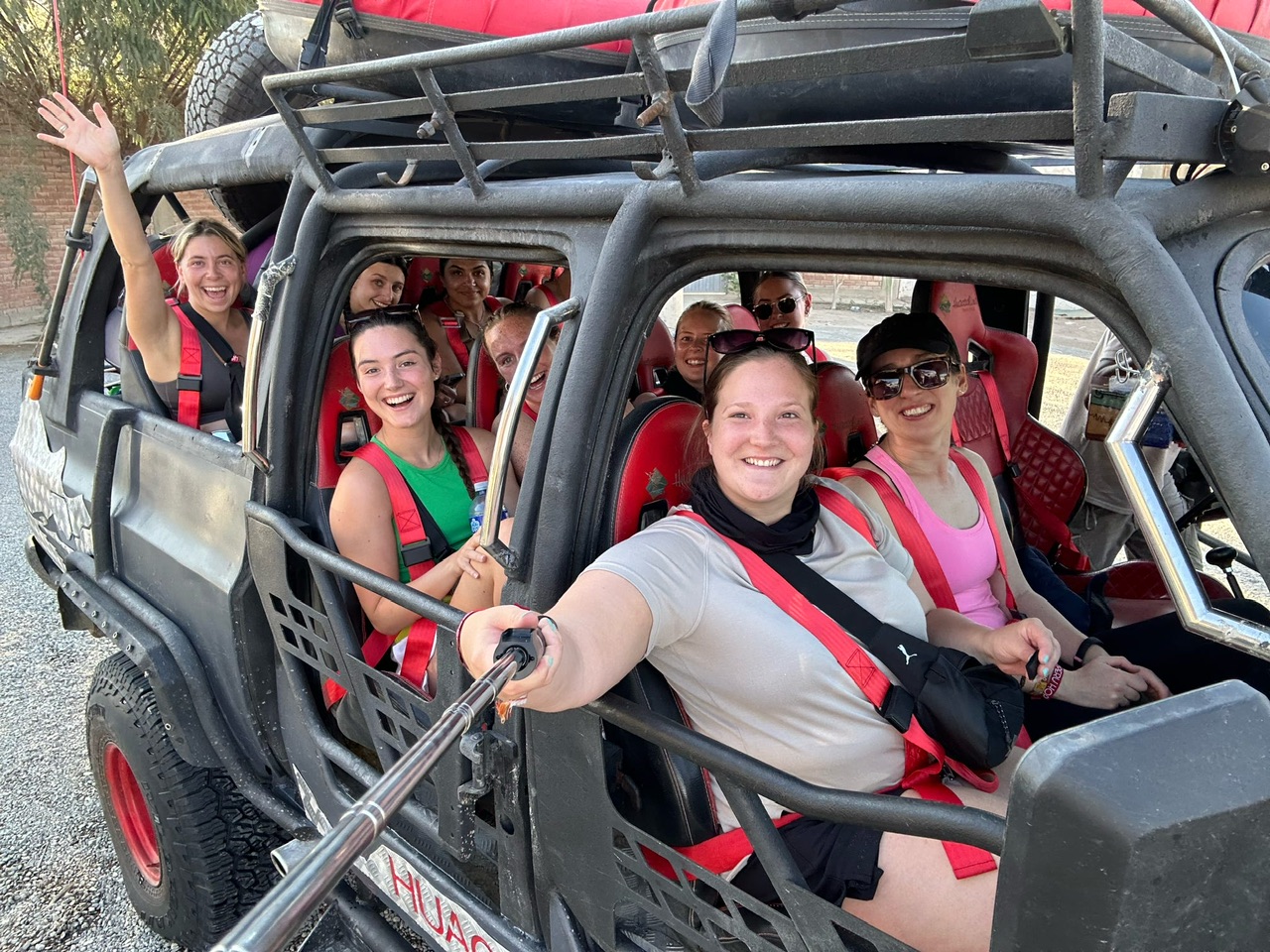 9 young women smiling in a dune buggy, woman in right front seat is holding selfie stick to capture photo.