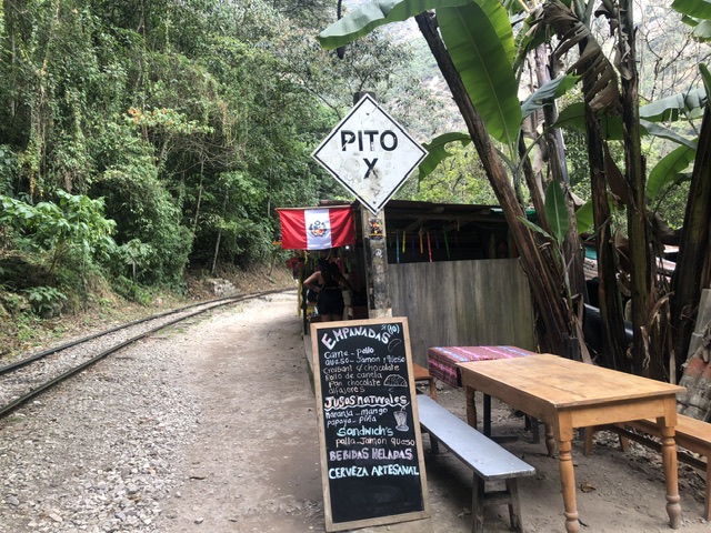 On the left is a trainline passing by a jungle On the right is a blackboard listing Emanadas for sale and a small wooden hut with a peru flag, sign saying "PITO X" and two small wooden tables with wooden benches down each side.