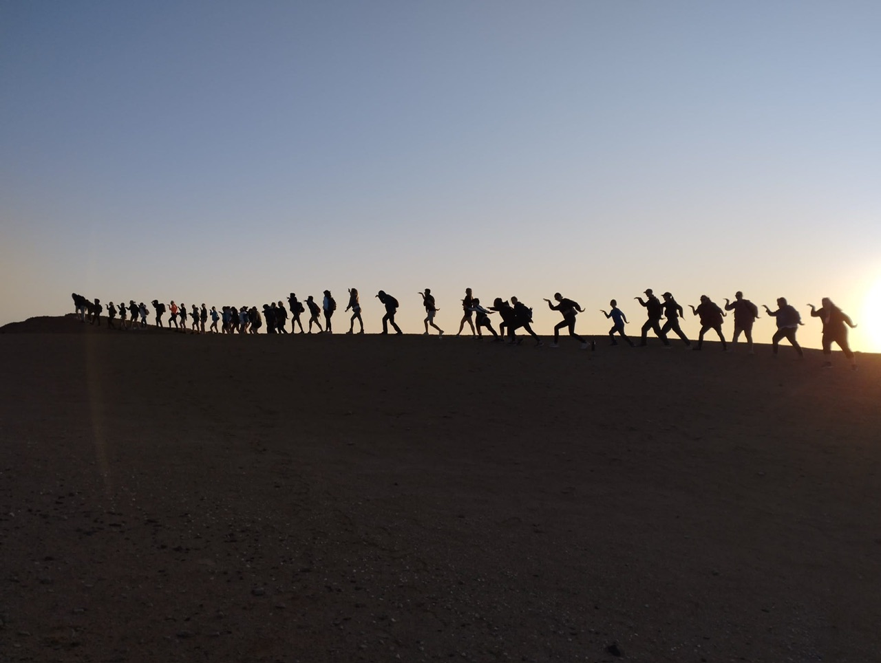 The silhouettes of around 40 people standing sideways facing the right in an egyptian walking position at the top of a sand dune with the sun setting behind.