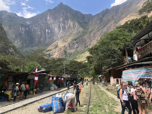 Shops and restaurants alongside traintracks at Hidroeléctrica Peru with walkers standing around in groups preparing to walk to Machu Picchu. Mountain ranges are in the background.