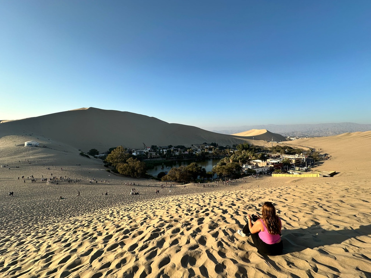 Young woman wearing black leggings and a pink singlet sits on a sand dune overlooking the Huacachina Oasis in Peru.