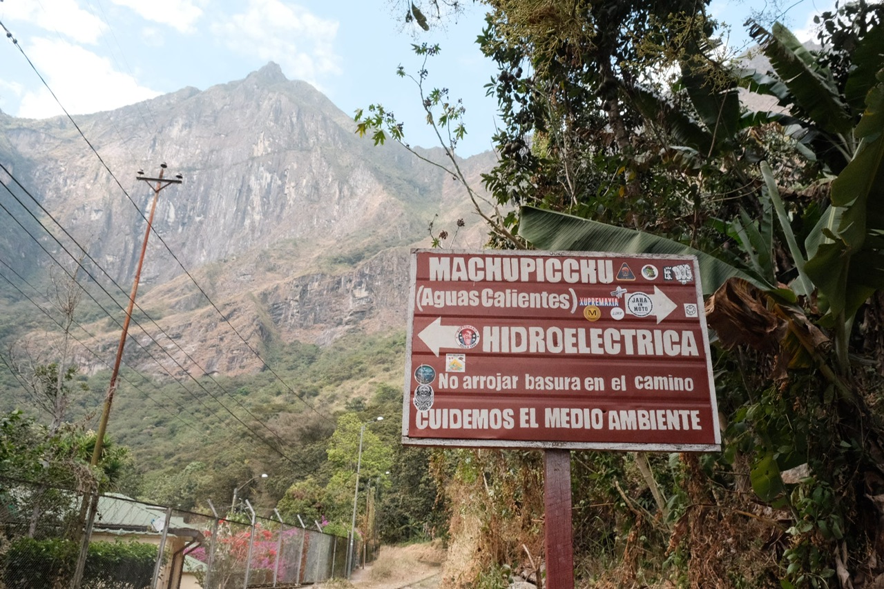 Brown sign to Machu Picchu and Aguas Calientes at Hydroelectrica in Peru, with mountains in background.