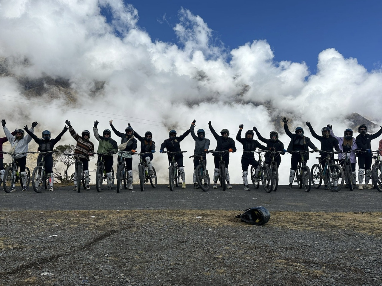 A group of 18 young women wearing helmets and protective gear stand with mountain bikes and their arms in the air. Clouds and mountains are in the background.