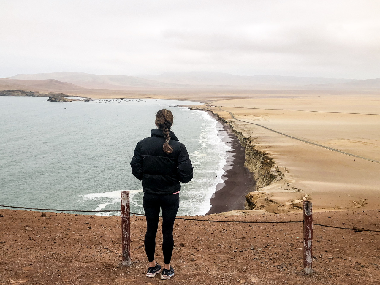 A young woman with long brunette hair in a plat stands facing away from the camera looking over the ocean from a viewpoint in Paracas National Reserve in Peru. She is wearing black leggings and a black puffer jacket and has her hands in her pockets.P