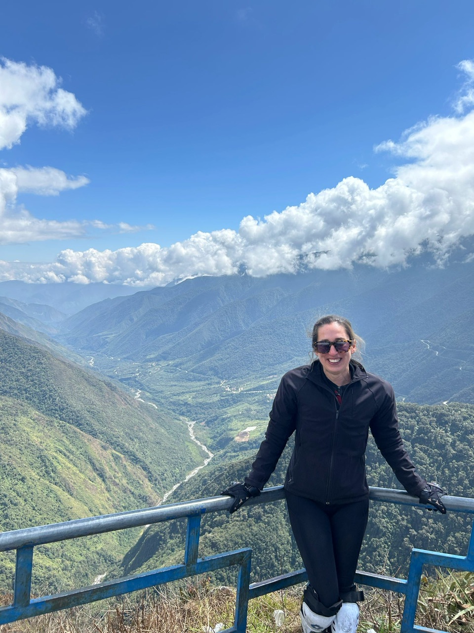 A young brunette woman wearing tortoiseshell sunglasses, black outdoor clothing, protective white kneepads and bike gloves stands smiling at the camera with the andes mountains in the background.