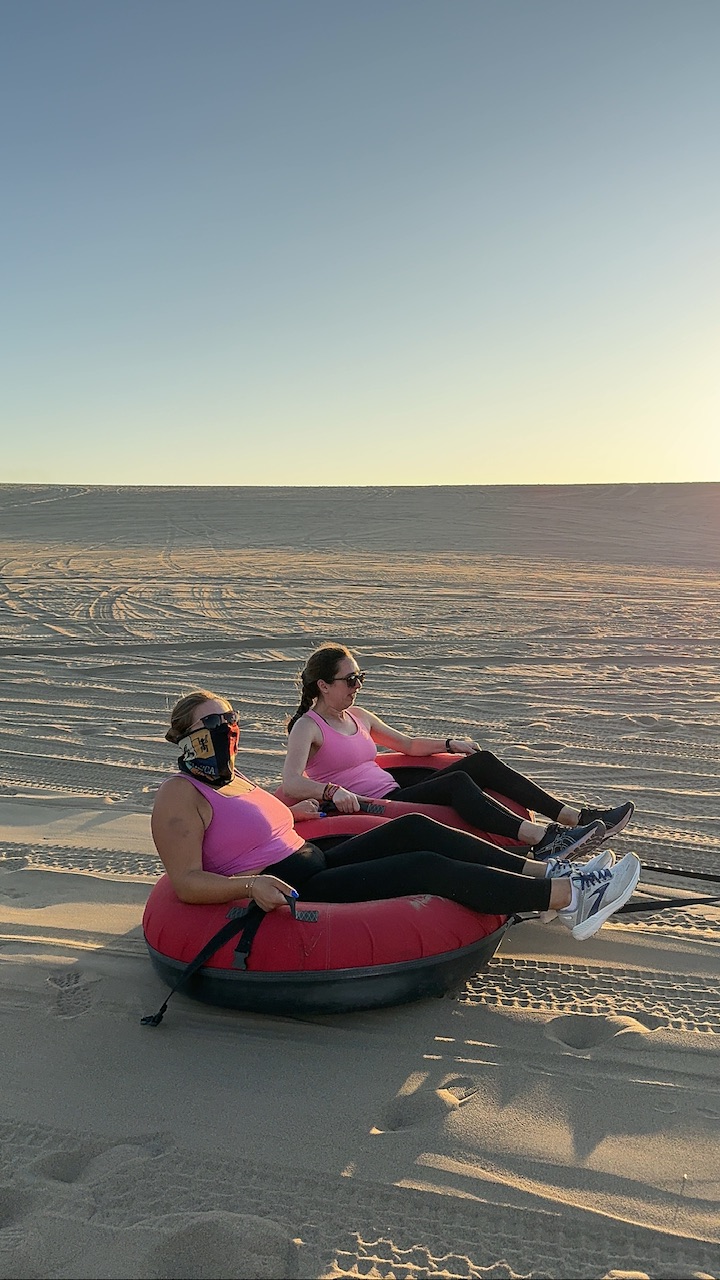 Two young brunette women wearing pink singlets, sunglasses and black leggings are towed on inflatable biscuits in the Huacachina desert.