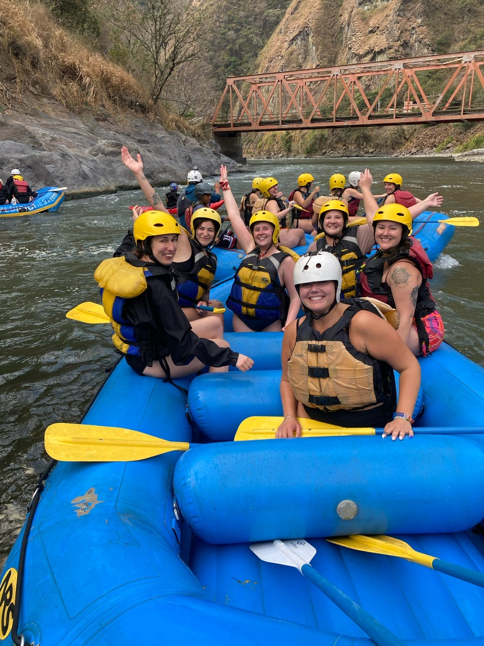 Six young women wearing helmets and lifejackets over swimwear sit towards the back of a blue white water raft holding oars and smiling on a river. In the background is another raft full of people and a brown footbridge over the river.