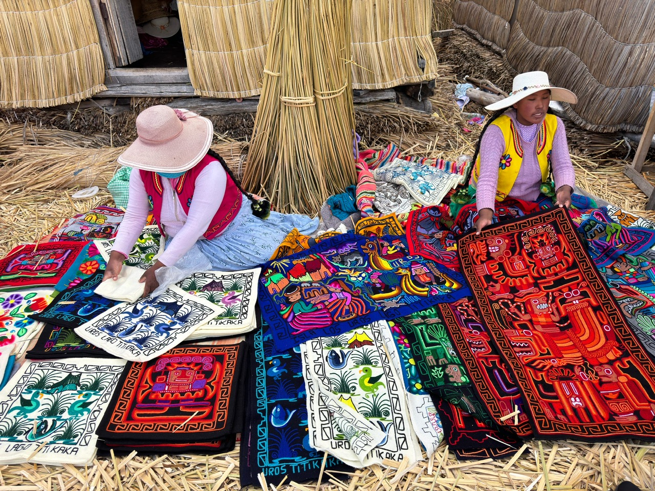 two women wearing traditional Peruvian clothing including wide brimmed hats sit on the ground displaying cushion covers and fabric hangings for sale. A thatched house is in the background and the ground is covered in flax.