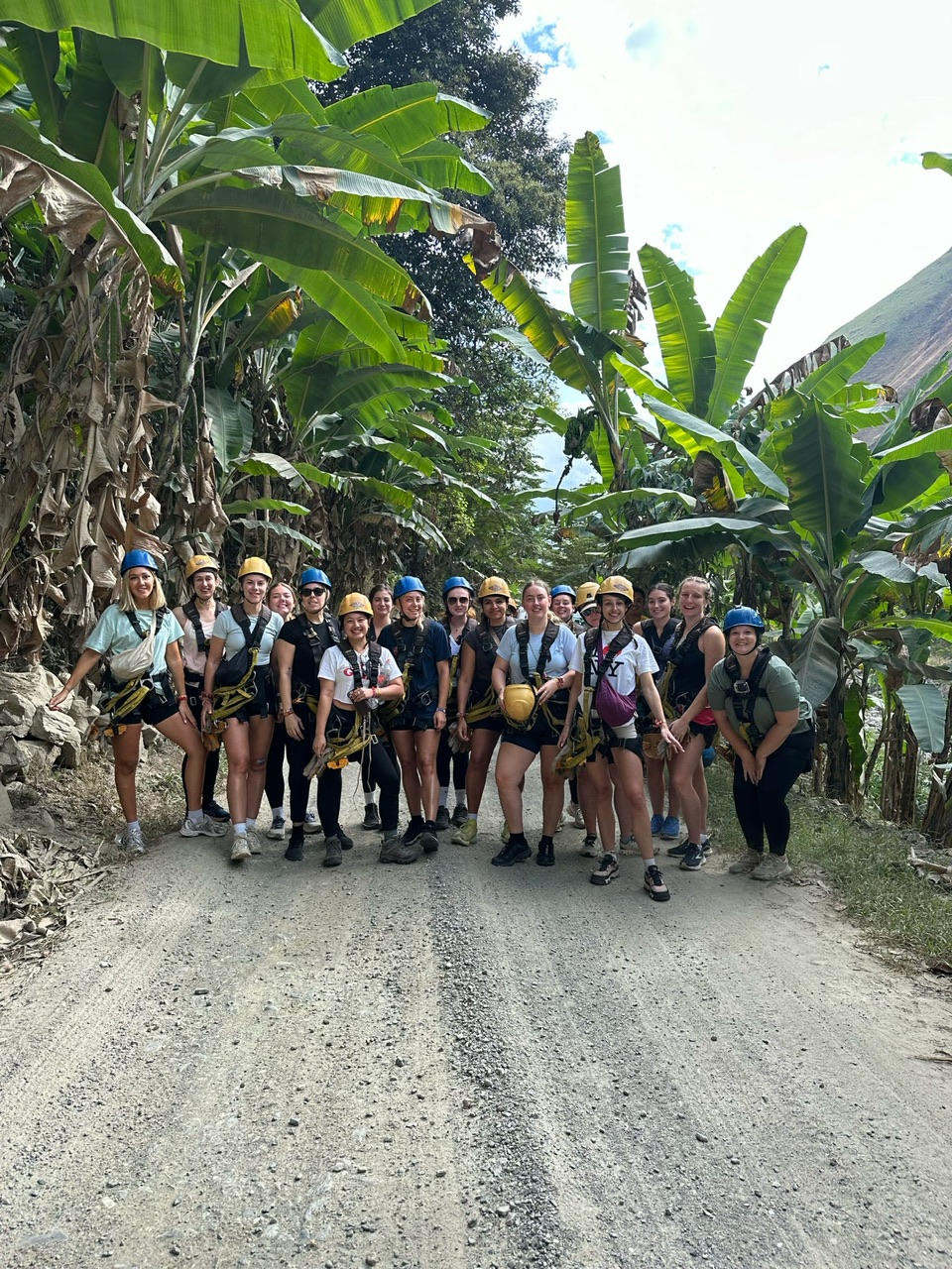 Group of 17 young women on Plotpackers tour of Peru wearing yellow and blue helmets and ziplining gear on a gravel road through the jungle.