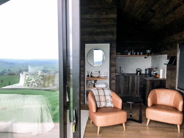 Looking into Rua Hiwi Hut through glass sliding doors. Kitchen is on right with two leather arm chairs in front, bathroom vanity is visible through open sliding door and bed is on the left of the room.